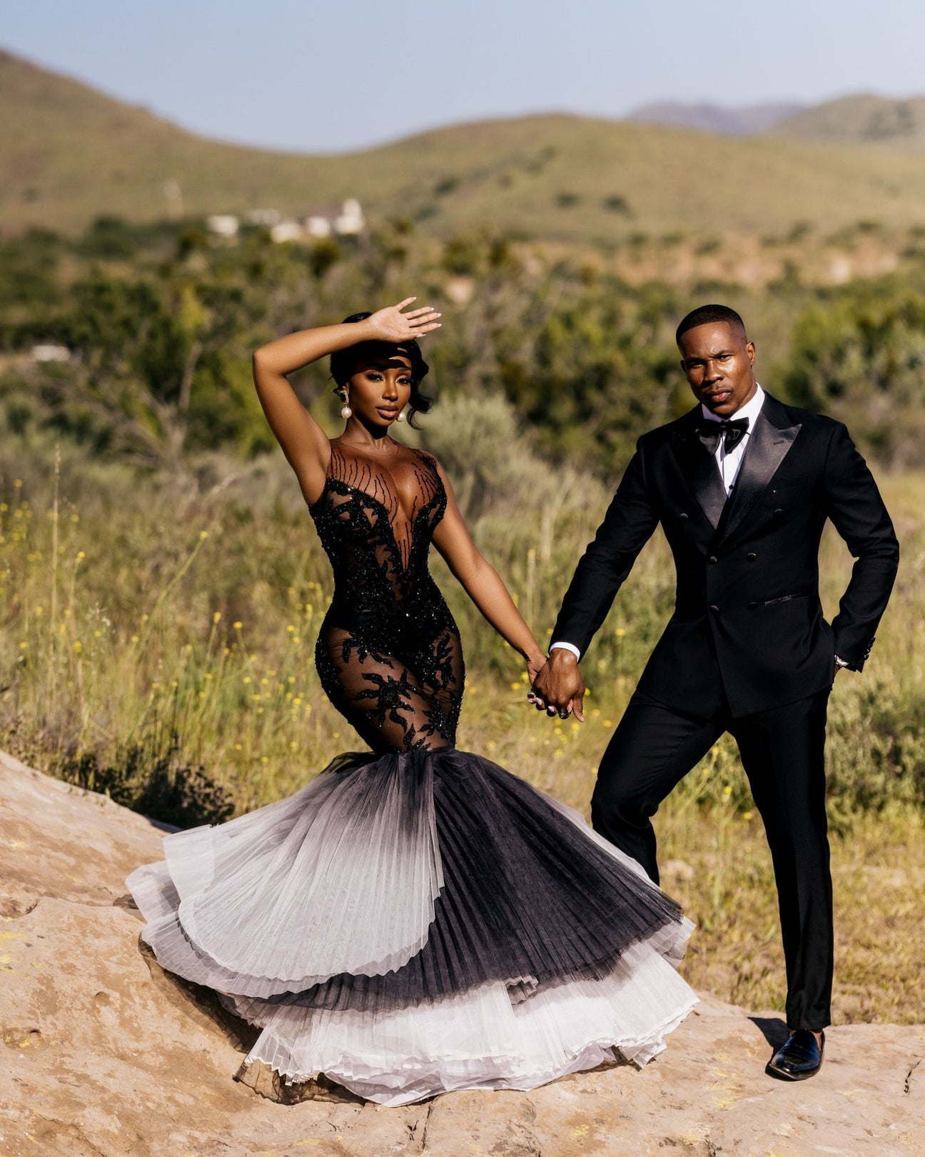 Elegant African Couple in Formal Attire - Stylish woman in black lace mermaid dress with flowing skirt and man in sleek black tuxedo pose outdoors amidst lush green foliage and distant mountains.