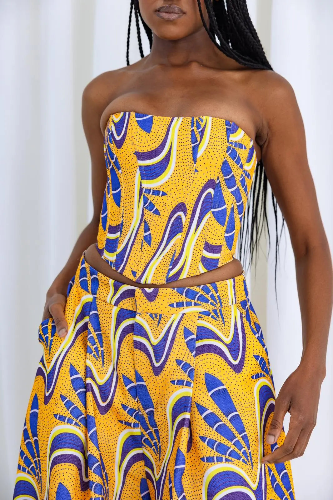 Vibrant African-inspired printed corset top featuring a bold, colorful pattern in shades of yellow and blue. The strapless design showcases the model's shoulders and neckline, creating a stylish and eye-catching look. This piece is part of the Claude Kameni fashion collection, which celebrates the rich heritage of Nigerian fabrics and African artistry.