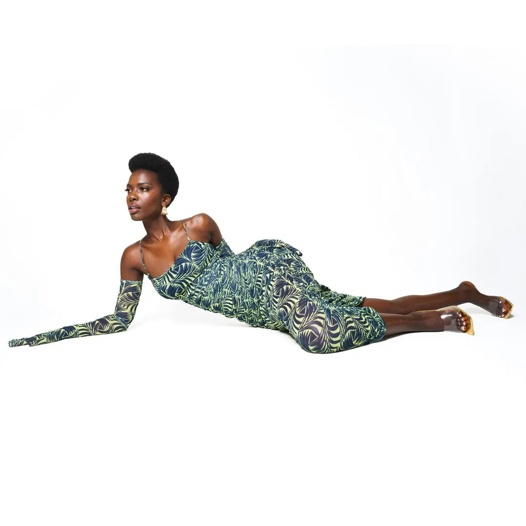 Elegant African-inspired dress with bold tropical print design, featuring a vibrant green and purple color palette. The model is lying on the floor, showcasing the dress's flowing silhouette and eye-catching pattern.