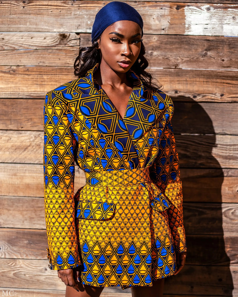 Stylish African print blazer with geometric patterns in vibrant yellow and blue colors, worn by a fashionable model against a wooden background.