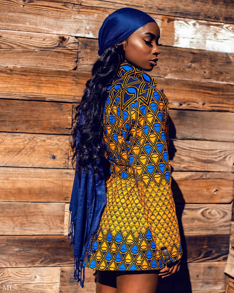 A vibrant African-inspired blazer with a bold print featuring a geometric pattern in shades of blue and yellow, worn by a young woman with dark curly hair against a rustic wooden background.