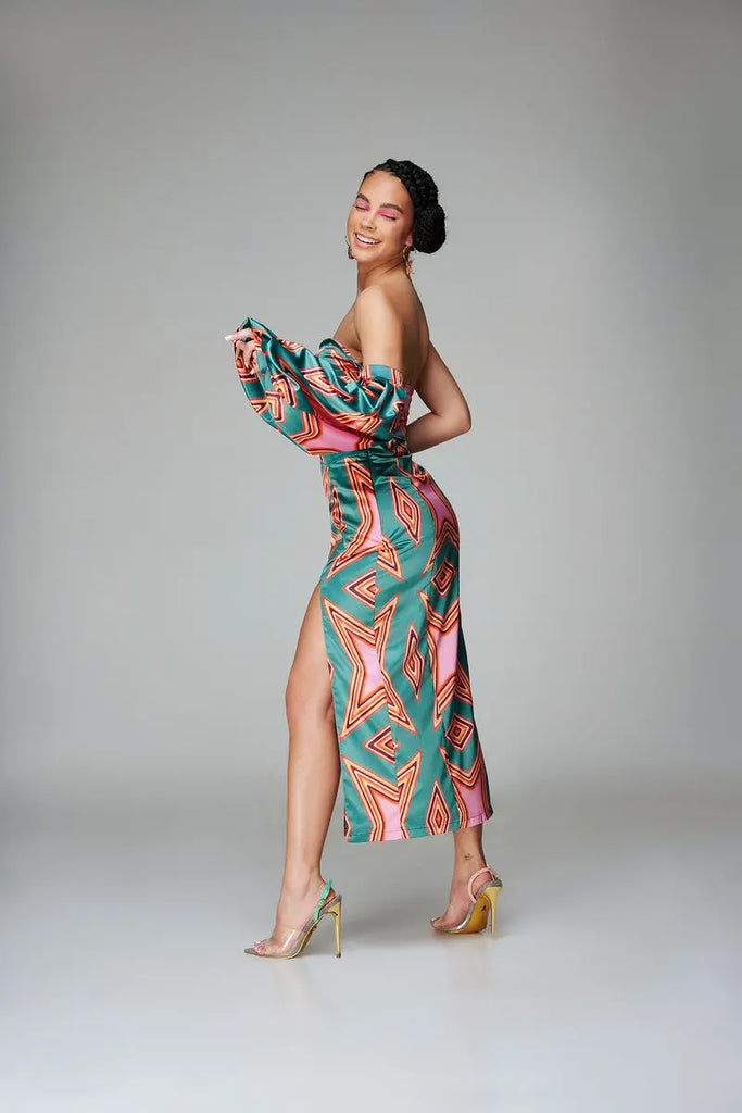 Vibrant Geometric-Print African Satin Midi-Dress
The image depicts a model wearing a beautifully-patterned, off-the-shoulder satin midi-dress from the African fashion brand Claude Kameni. The colorful geometric print design features a high slit skirt and sleeves, creating a bold, trendy look.
