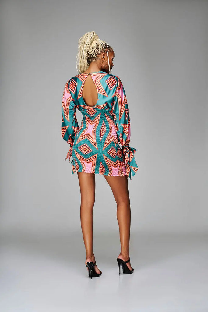 Vibrant African print mini dress with bold geometric patterns and a low-cut back detail, worn by a blonde female model posing on a plain background.