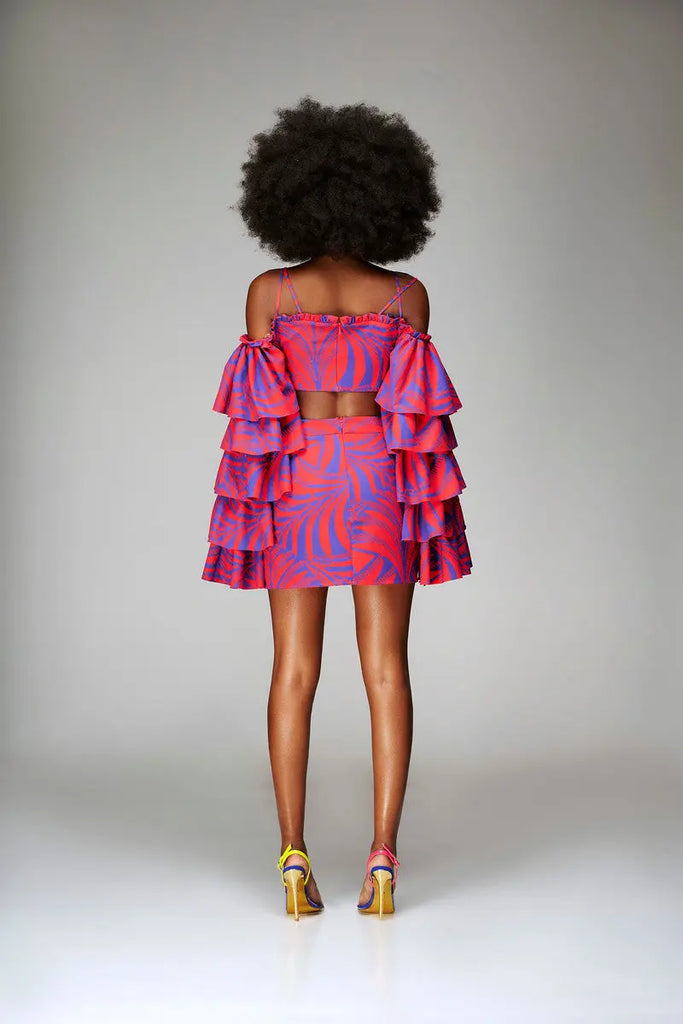 Vibrant African print ruffled top with striking pink and blue pattern, worn by a woman with natural curly hair, standing in a simple studio setting.